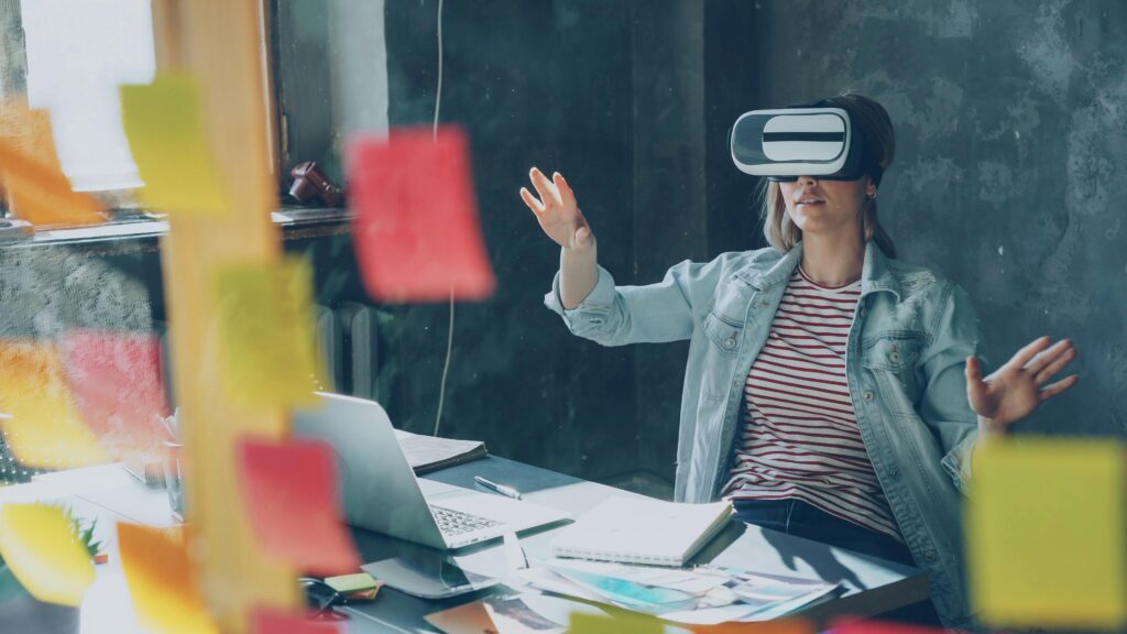 Persona usando gafas de realidad virtual mientras trabaja en un entorno de oficina creativa con notas adhesivas, portátil, cuadernos y materiales de planificación sobre la mesa, representando innovación y estrategia digital para el curso Programa Avanzado en Proyectos, Estrategia e Innovación Digital - ADGD27