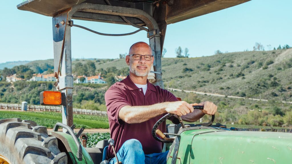 Agricultor con gafas conduciendo un tractor verde en una explotación agrícola de montaña, con paisaje rural de viñedos y bosques al fondo.