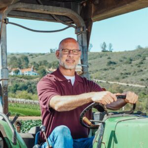 Agricultor con gafas conduciendo un tractor verde en una explotación agrícola de montaña, con paisaje rural de viñedos y bosques al fondo.